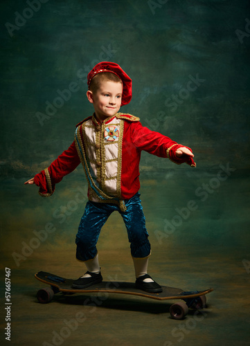 Smiling little boy dressed up as medieval little prince and pageboy skateboarding over dark vintage style background. Action, leisure activities, emotions, sport concept