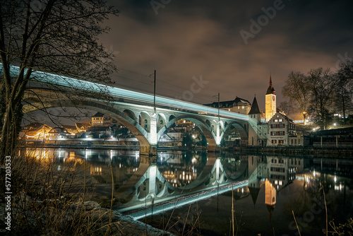 Nachtaufnahme der historischen Stadt Bremgarten mit der Eisenbahnbrücke im Vordergrund. Die Brücke reflektiert sich in der spiegelglatten Reuss.