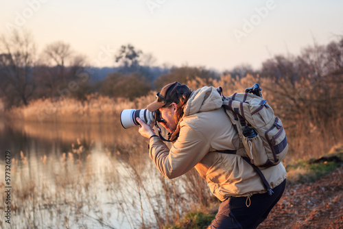 Photographer with camera, telephoto lens and backpack taking picture at lake during sunset. Hobby and outdoor leisure activity