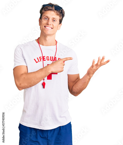 Young handsome man wearing lifeguard t shirt and whistle amazed and smiling to the camera while presenting with hand and pointing with finger.