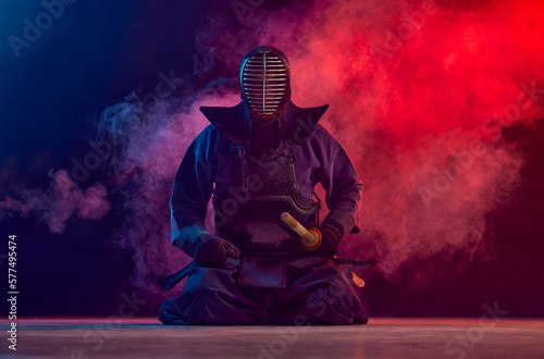 Man, professional kendo athlete, combat sportsman posing with shinai sword against gradient dark background in neon light with smoke. Concept of martial arts, sport, Japanese culture, action and