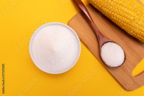 Natural sweetener Erythritol, produced by fermentation from corn in ceramic bowl, wooden spoon, corncob on yellow orange background. Sugar substitute. Horizontal plane, top view.