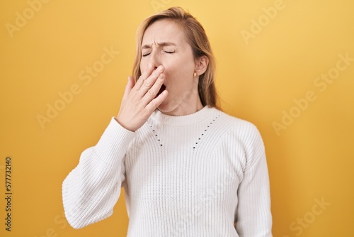 Young caucasian woman wearing white sweater over yellow background bored yawning tired covering mouth with hand. restless and sleepiness.