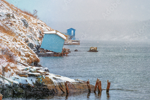Two vibrant blue colored wooden fishing sheds at the bottom of a hill. One boat house is tipping towards the ocean. There's snow on the grassy hill and it's snowing heavily coming in the cove. 
