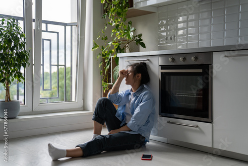 Unhappy Asian girl sitting on floor in kitchen feeling depressed after social media usage, being ignored by boyfriend, dealing with rejection in love. Breakup depression concept