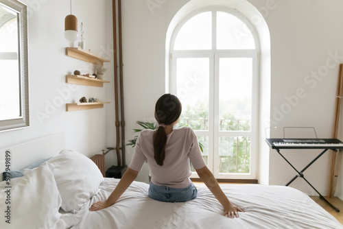 Back view of relaxed young woman sitting on bed with white linen, looking forward at window, enjoying relaxation in bedroom, hotel room, city view, comfort at cozy home