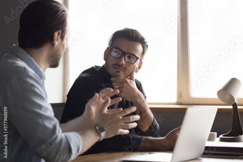 Focused stylish millennial businessman consulting expert, listening to professional advice from boss, coworker, mentor. Business colleagues men meeting, talking at office work table