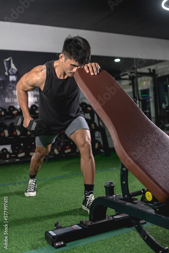 A determined man does one-arm dumbbell rows while leaning on an incline bench. Back exercise variation. Weight and resistance training at the gym.