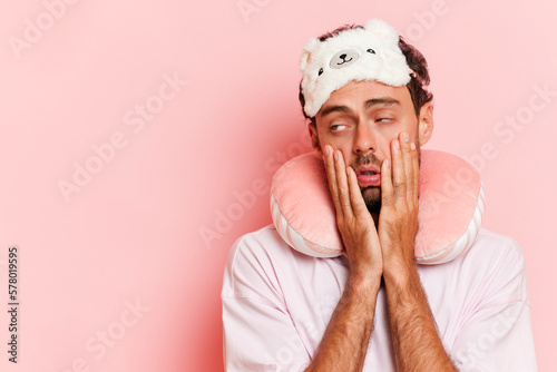 Horizontal shot of tired unshaven caucasian man wears sleeping mask neck pillow looks exhausted while traveling isolated over pink background. Transport, tourism, road trip concept. 
