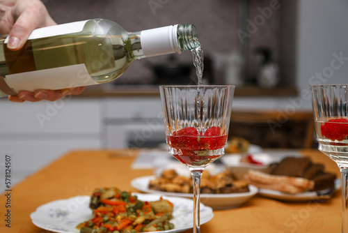 Woman pouring white wine from bottle in a wine glass with frozen strawberry at a dinner at home