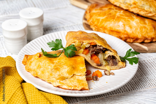 Baked meat turnovers or pies, or empanadas, or cornish pasty with filling, beef, carrot, and potato, white plate.