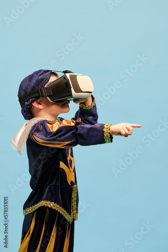 One little boy in costume of medieval pageboy, little prince wearing VR headset standing over blue background. Emotions, virtual reality, games, dreams concept