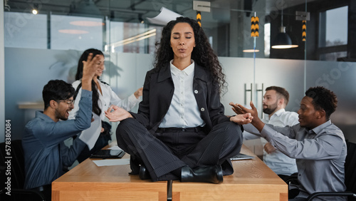 Calm businesswoman professional taking break meditating sitting at table in office meeting ignoring loud colleagues discussing brainstorming argue mindful healthy female boss doing yoga no stress zen