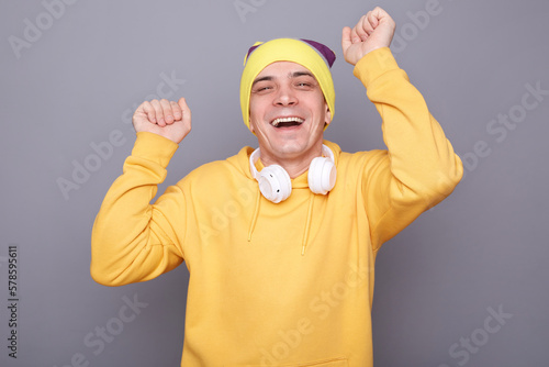 Image of festive happy cheerful man wearing beanie hat and yellow casual hoodie standing isolated over gray background, posing with raised arms, dancing and laughing, celebrating.