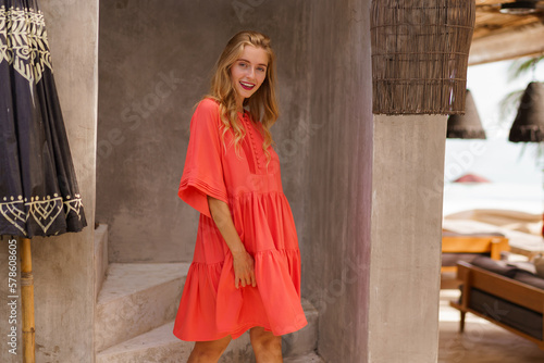 Pretty smiling blond woman in summer red dress poaing ib beautiful tropical resort. Vacation mood.