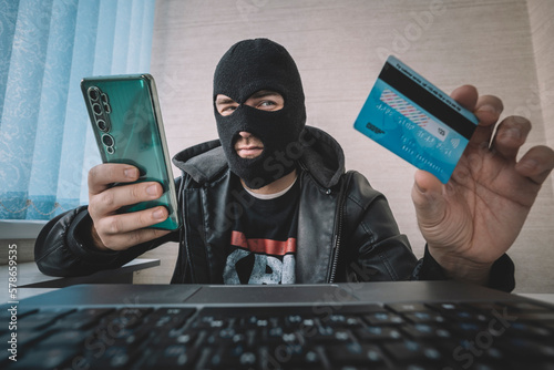 Male hacker in a robber mask uses phone, credit card and laptop in some fraudulent scheme. Cyber thief stole the personal data and credit card information. Hacker uses malware to steal user's money