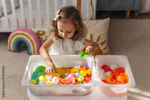 A little girl playing with colored rice and Easter eggs in sensory bin. Easter sensory bin with bright rice and eggs, bunny, carrot. Sensory play and holidays activity for kids. Happy Easter concept.