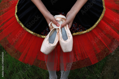 Looking down from above at the waist of a ballerina wearing a red and black tutu with gold colored trim holding her pink ballet pointe shoes in both hands.