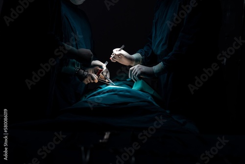 Surgical team performing surgery to patient in sterile operating room. In a surgery room lit by a lamp, a professional and confident surgical team provides medical care to an unconscious patient.