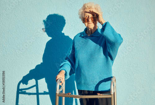 Portrait, shadow and disability with a senior woman on a blue wall background while holding a mobility walker outdoor. Health, handicap and fashion with a mature female standing outside alone