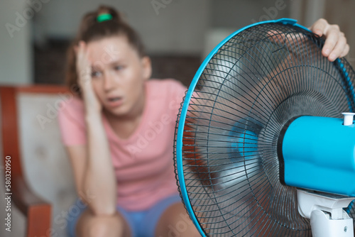 Young woman using electric fan at home in living room, sitting on couch cooling off during hot weather, suffering from heat, high temperature