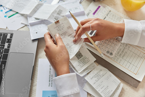 Unrecognizable woman holds receipts pencil uses laptop plans budget manages expenses calculates financial reports checks internet banking service poses at desktop with papers around. View from above