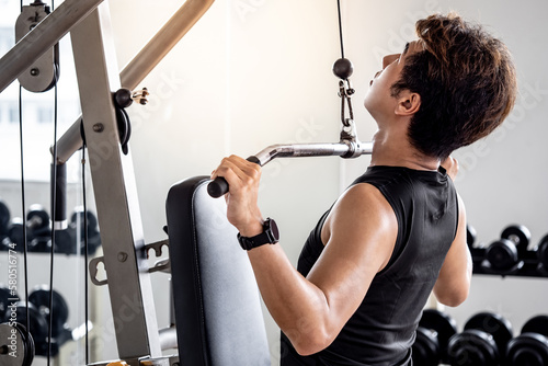 Asian sport man with well trained body in black sportswear pulling overhead bar doing exercise training with lat pulldown machine in fitness gym. Muscular build and physical training concept.