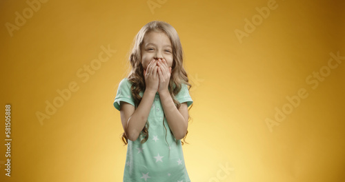 Studio shot of little caucasian girl is covering her mouth with a hand with astonished face expression, isolated over yellow background.