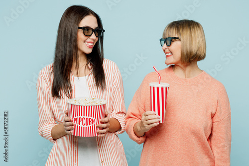Smiling elder parent mom with young adult daughter two women together in 3d glasses watch movie film hold bucket of popcorn cup of soda pop in cinema isolated on plain blue background studio portrait.