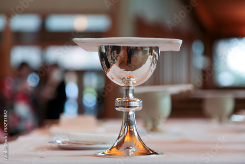 A shining silver communion chalice containing wine, a sacrament, is covered with a chalice pall is resting on a table in an anglican church