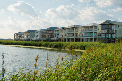 Row of houses under the bright sky with clouds with lagoon waterfront at Destin, Florida. There is a lagoon at the front with tall grasses near the shore and a boardwalk pavilion on the right.