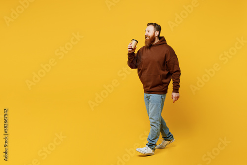 Full body young redhead caucasian man wear brown hoody casual clothes hold takeaway delivery craft paper brown cup coffee to go isolated on plain yellow background studio portrait. Lifestyle concept.