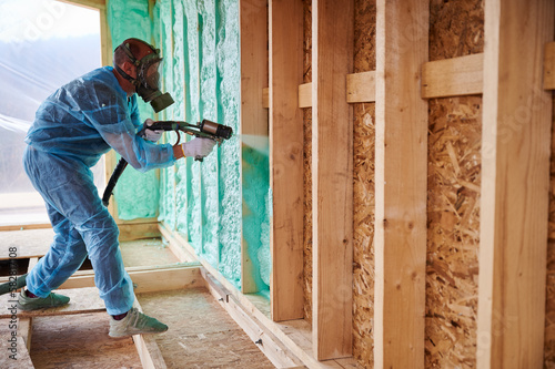 Male builder insulating wooden frame house. Man worker spraying polyurethane foam inside of future cottage, using plural component gun. Construction and insulation concept.