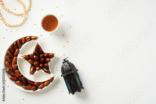 Ramadan dates in crescent moon plate, cup of tea, rosary, oriental lantern on white background. Ramadan Kareem concept.