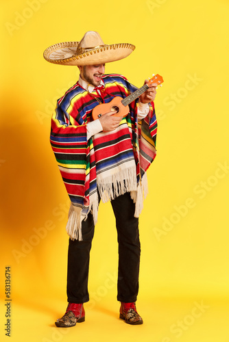 Portrait of young handsome man in colorful clothes, poncho and sombrero playing guitar, posing against yellow studio background. Concept of mexican traditions, fun, celebration, festival, emotions