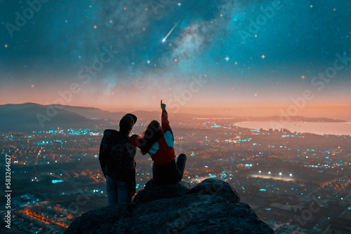 silhouettes of a couple sitting on the top of the mountain looking and pointing out at shooting star and milky way over the city lights on the horizon 