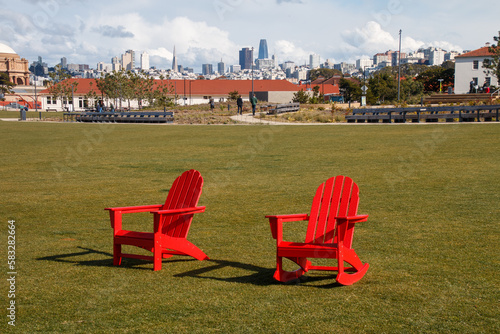 Two red chairs are sitting on green grass at The Presidio's Tunnel Top Park. In the background is the San Francisco cityscape. There is a blue sky with white clouds.