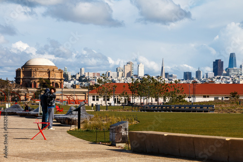 A view of the San Francisco skyline from the Tunnel Top park in The Presidio. Two people are standing to the side. There white clouds in a blue sky.