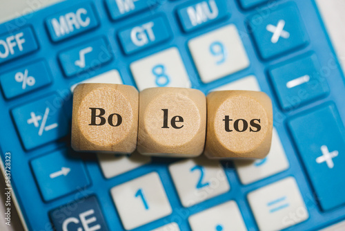 A palavra boletos em Português do Brasil separada por sílabas escritas em dados de madeira que estão sobre uma calculadora azul. Economia brasileira.