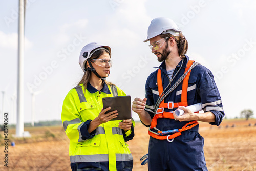 Caucasian engineer man discuss together with co-worker woman hold blueprint with tablet stay in front of row of windmill or wind turbine, wind turbine generate electricity, Environmentally friendly