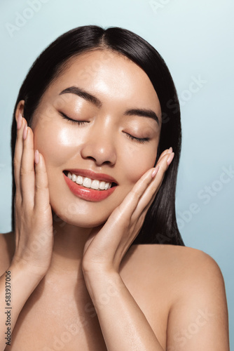 Vertical shot of smiling korean girl washes her face, applied skincare routine, facial cream with pleased face, touches her soft skin, stands topless against blue background