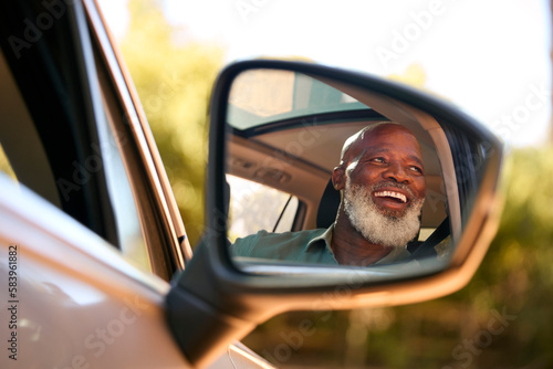 Smiling Senior Male Driver Reflected In Wing Mirror Of Car Enjoying Day Trip Out Driving