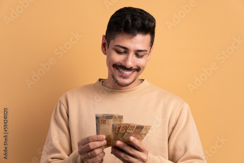 cheerful brazilian man with brazil banknotes currency in beige background. business, loan, pay, wealth concept. 