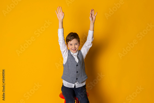 A happy little boy in gray vest and white shirt, neatly dressed, laughing and keeping his hands upwards. Isolated on yellow background.