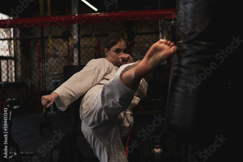 A girl in a kimono exercises with a punching bag in the gym while learning karate martial arts