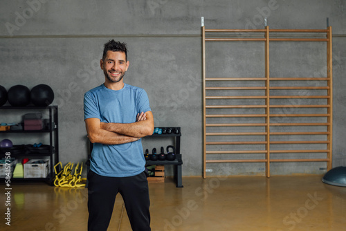 Portrait of Confident smiling hispanic man looking at camera in sport club. Fitness instructor standing arms crossed at gym. Real people, job occupation concept. Copy-space, horizontal