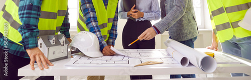 Construction team at a meeting. Group of young architects and builders discussing construction plans while standing around office table with paper blueprints and safety hard hats. Banner background