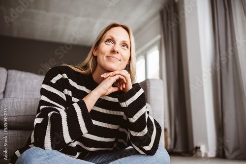 Beautiful Blonde Woman in her 30s Sitting on the Floor in Front of Sofa and Smiling at Something Off-Camera.