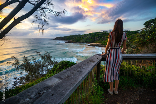 A girl in dress walks along the boardwalk and enjoys the view of the stunning first and second bay Coolum and big waves on Pacific Ocean. Stunning panorama of Sunshine Coast, Queensland, Australia