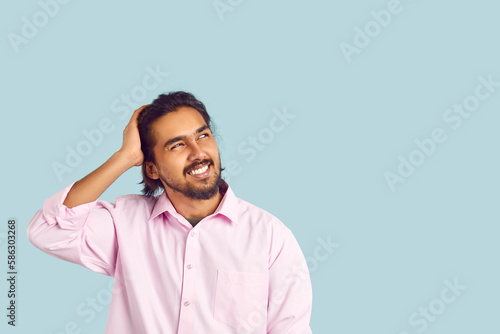 Man with funny facial expression is trying to remember what he forgot by looking at copy space. Portrait on pastel light blue background of confused Indian young man who doubts or made mistake.